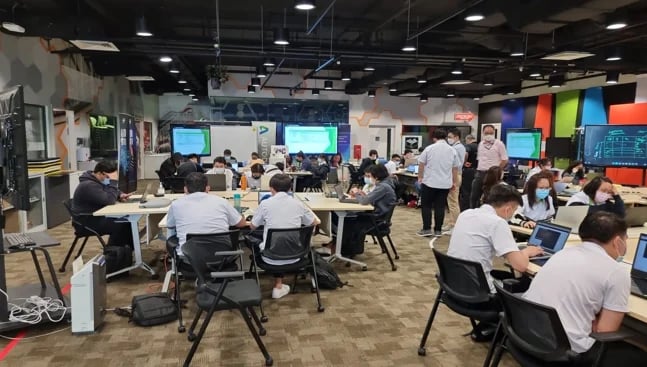Busy open-plan tech workspace with people seated at tables with computers and laptops, bright overhead lighting, and colorful wall displays