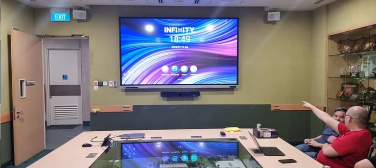Modern conference room with wall-mounted display showing blue and purple graphics, table with laptops, and person in red shirt seated at desk
